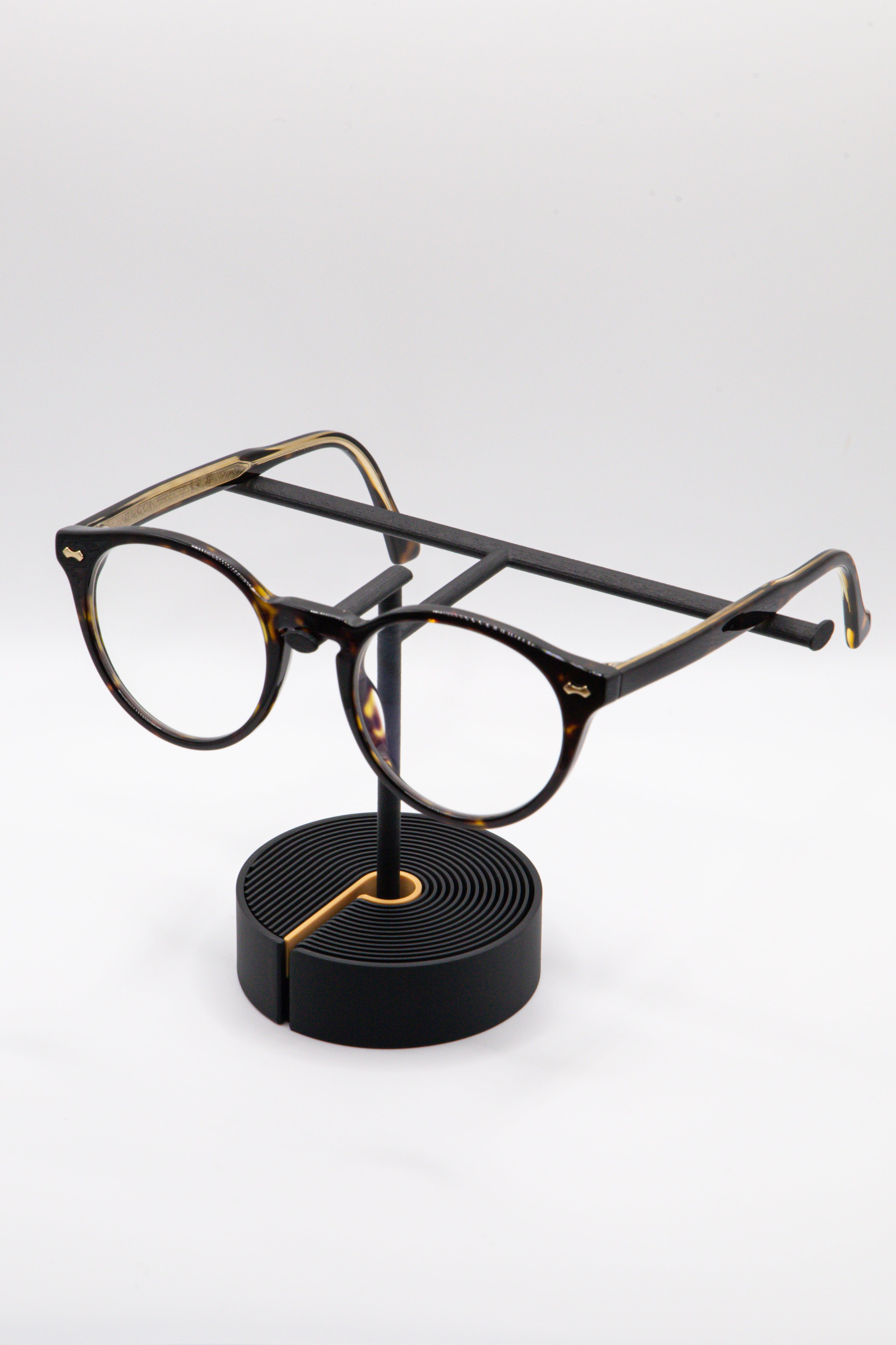 Brillenhalter PureStand in Schwarz matt mit runder schwarzer Base, goldenem Einelegeteil und Brille aufgesetzt.