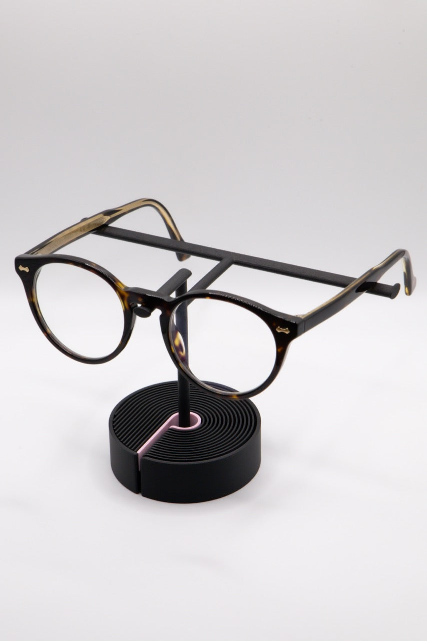 Brillenhalter PureStand in Schwarz matt mit runder schwarzer Base, pinkem Einelegeteil und Brille aufgesetzt.