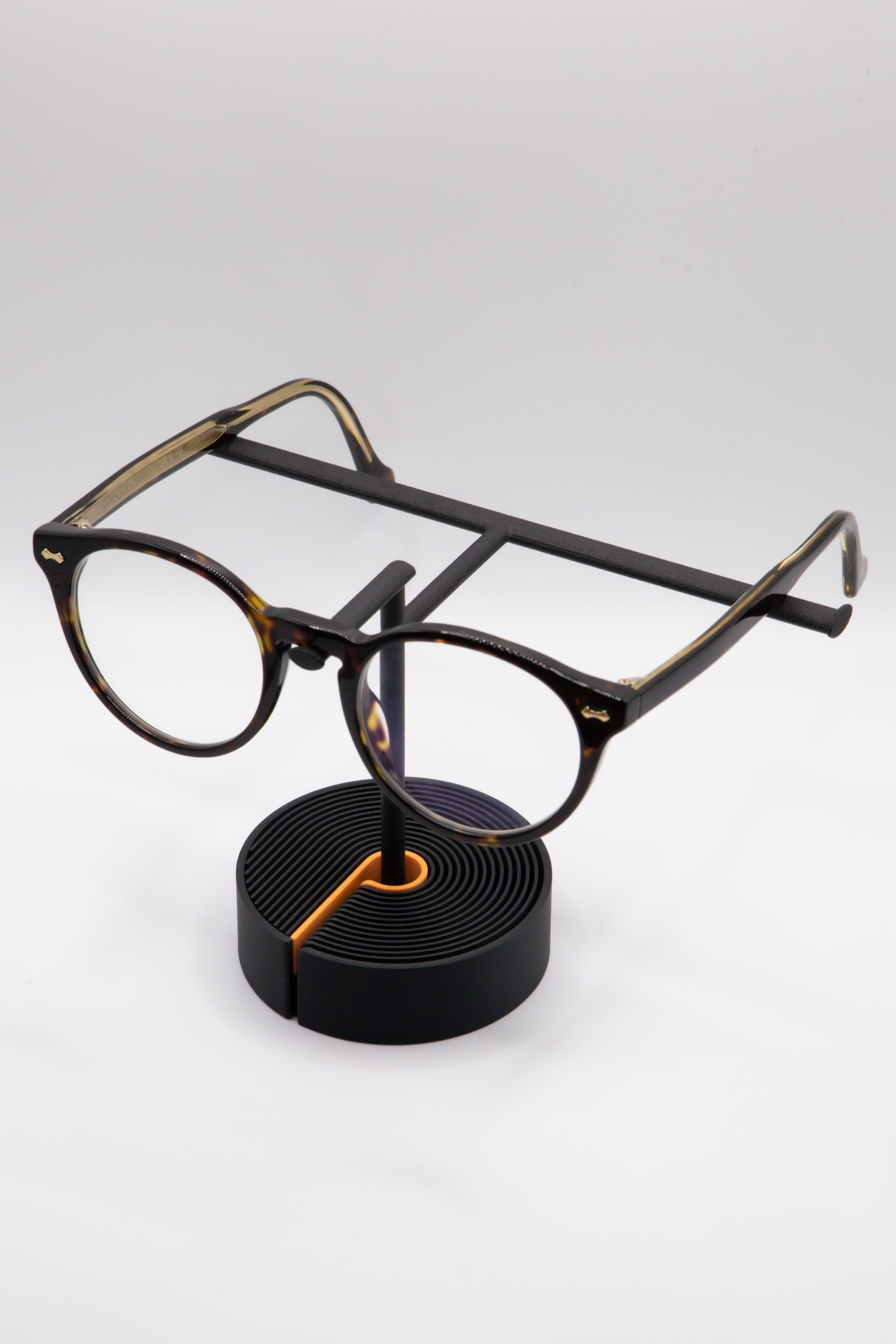 Brillenhalter PureStand in Schwarz matt mit runder schwarzer Base, orangenem Einelegeteil und Brille aufgesetzt.